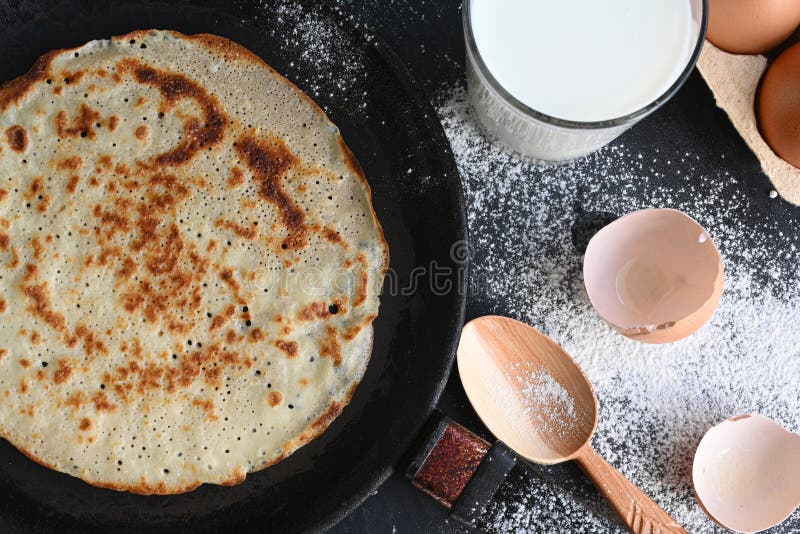 Hot Pancake in Black Pan on Black Table with Flour, Milk and Eggs Stock