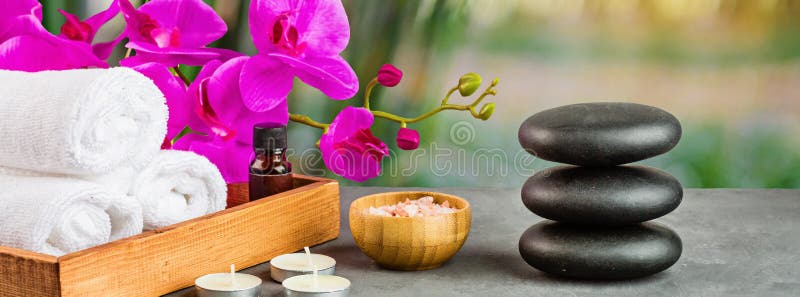 Hot spa stones set for massage treatment, orchid flower, towels, candles and sea salt on green background with bamboo. Elegant and
