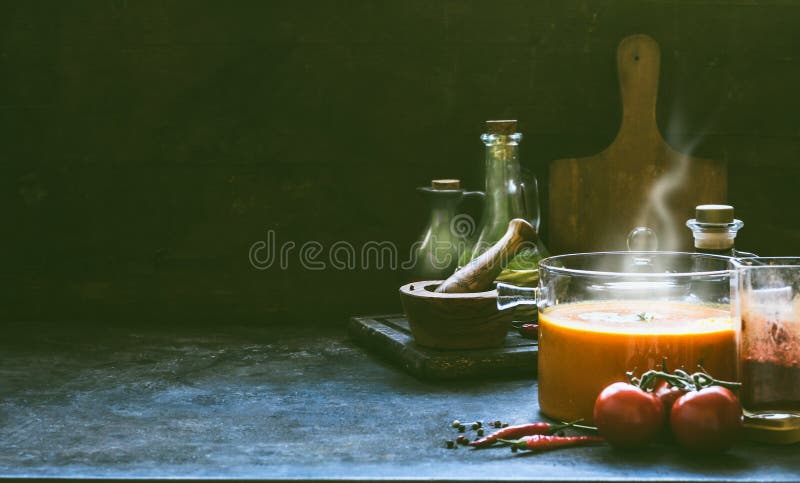 Hot tomato soup with steam in glass pot on rustic table at dark wall background with cooking ingredients. Kitchen scene , still