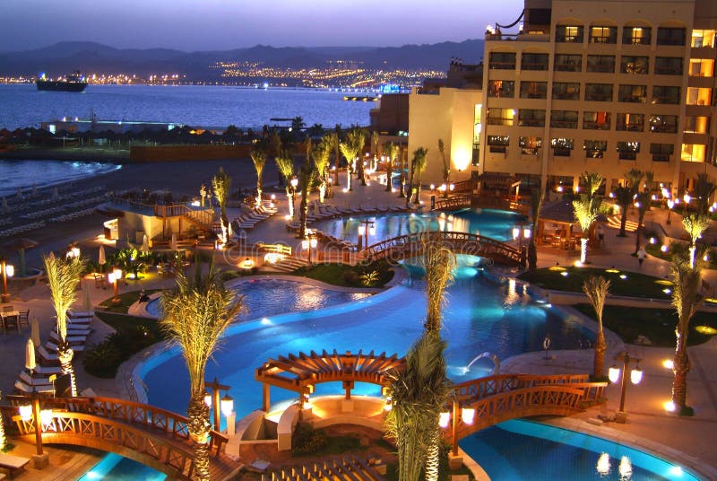 The luxurious Aqaba Jordan Hotel at twilight, illuminated swimming pools and lavish interior yard with city in the distance. Located on seaside. The luxurious Aqaba Jordan Hotel at twilight, illuminated swimming pools and lavish interior yard with city in the distance. Located on seaside.