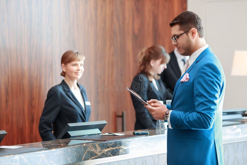 Looking for check-in information. Young handsome businessman in classical blue suit looks at his tablet device standing just in front of the hotel reception desk where young receptionists welcomes him with a smile. Looking for check-in information. Young handsome businessman in classical blue suit looks at his tablet device standing just in front of the hotel reception desk where young receptionists welcomes him with a smile