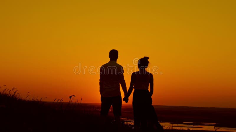 Houdend van paar bij zonsondergang - de jonge mens en de mooie meisjestribunes bij de zomerweide en het kijken aan zon, silhouett