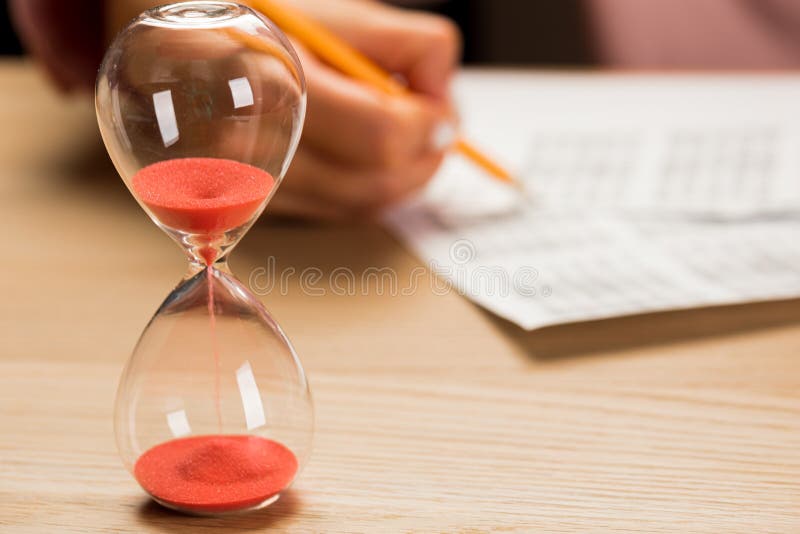 .Hourglass with time running out and student hand testing in exercise and passing exam carbon paper computer sheet with pencil in school test room
