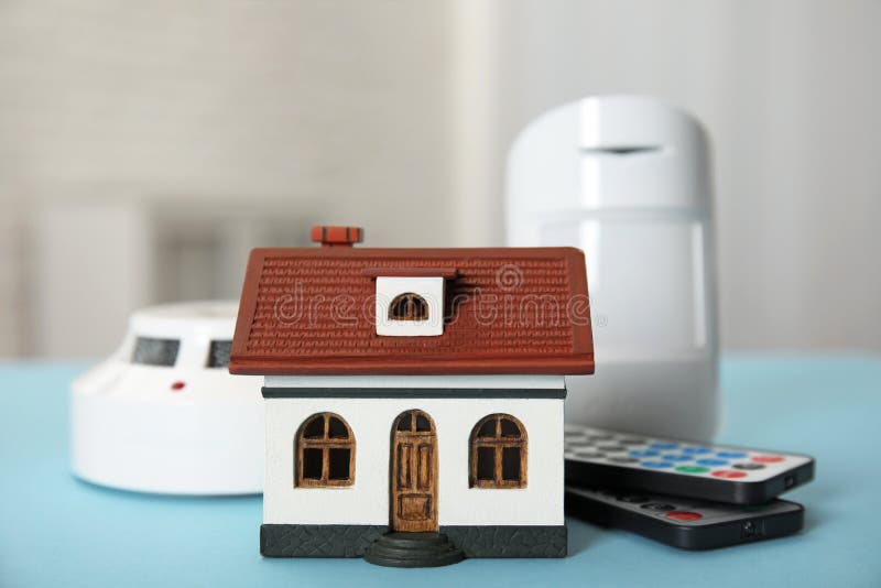 House model, remote controls, smoke and movement detectors on light blue table in room. Home security system