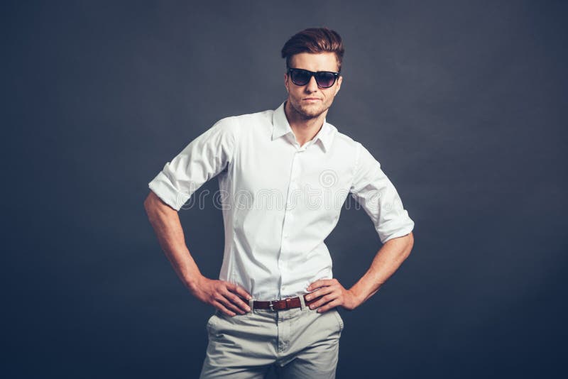 Confident young handsome man in sunglasses keeping arms akimbo and looking at camera while standing against grey background. Confident young handsome man in sunglasses keeping arms akimbo and looking at camera while standing against grey background