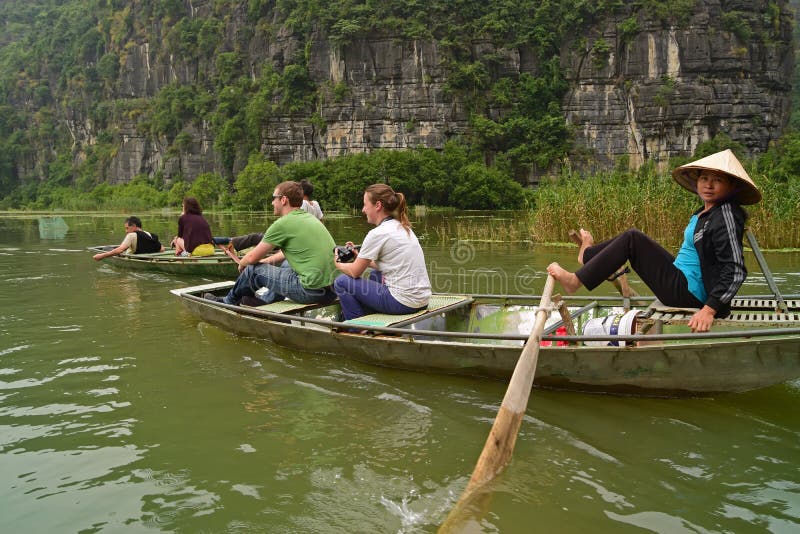 Tourists enjoy the ride & boat rower looking at the camera while rowing using feet through cliffs at Tam Coc, Vietnam