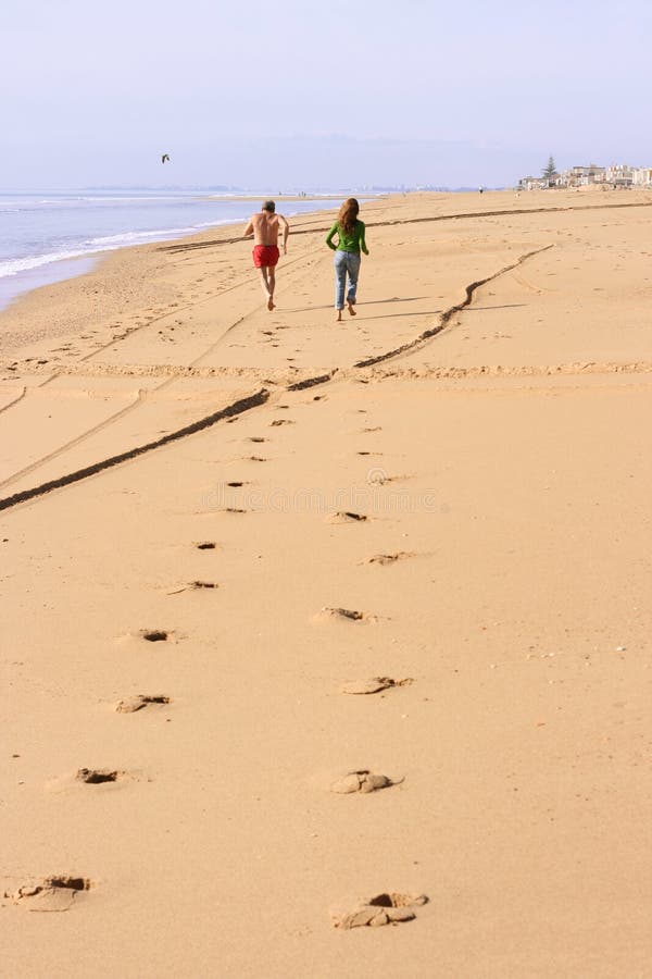 Footprints of a senior man in swimsuit and young woman with casual clothes running at the beach next to the water. The two subjects are at the end of the footprint way. A coastal town is visible at the end of the beach. Footprints of a senior man in swimsuit and young woman with casual clothes running at the beach next to the water. The two subjects are at the end of the footprint way. A coastal town is visible at the end of the beach.