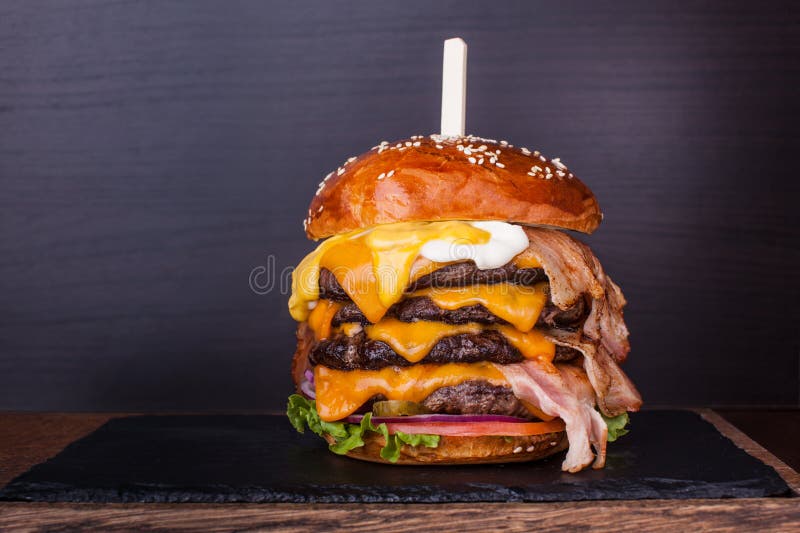 Huge burger with beef, bacon, cheese, fresh vegetables and sauces on plate over wooden background. For restaurant menu