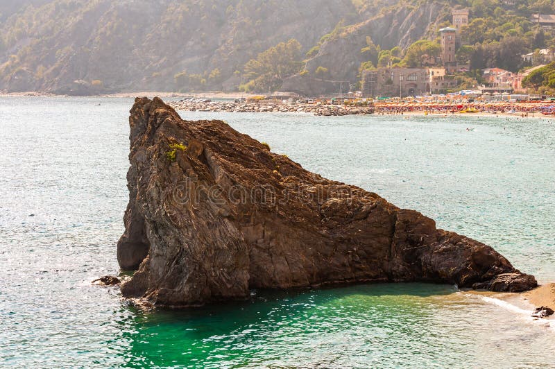 Huge triangle shape rock in the water on the beach with famous cityscape of Monterosso Al Mare and high green mountains on the