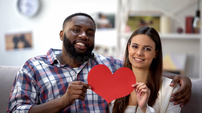 Hugging mixed-race couple showing paper heart to camera, sitting on sofa at home royalty free stock image Hugging mixed-race couple showing paper heart to camera, sitting on sofa at home royalty free stock image