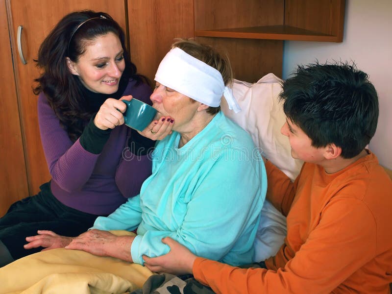 Daughter and grandson take care of sick grandmother (bandaged head) in bed, give her a cup of hot drink because she has a fever. Horizontal color photo. Daughter and grandson take care of sick grandmother (bandaged head) in bed, give her a cup of hot drink because she has a fever. Horizontal color photo.