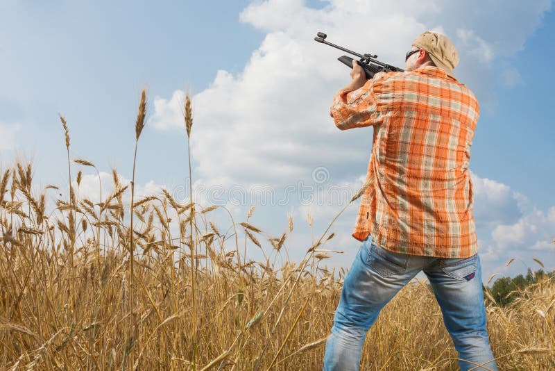Hunter in cap and sunglasses aiming a gun at field