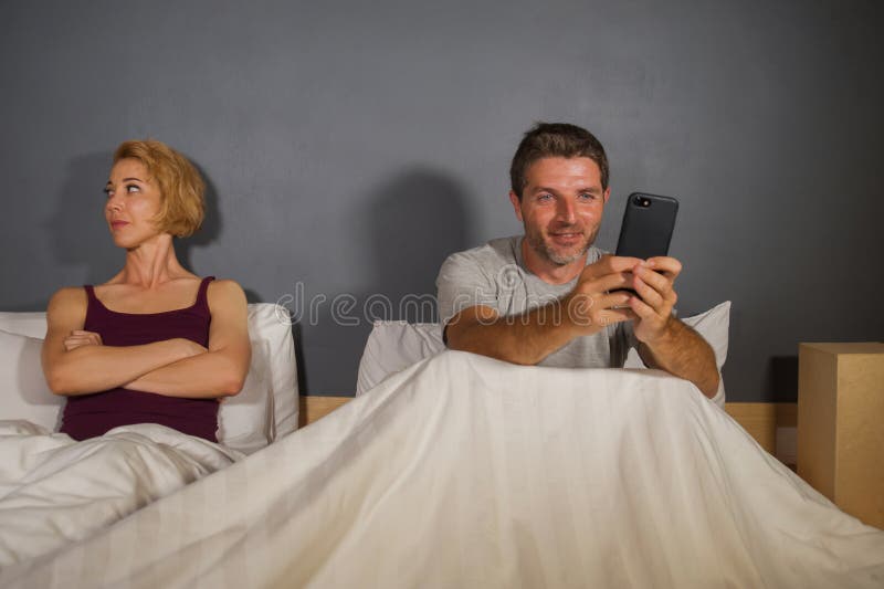 Husband or boyfriend using mobile phone in bed and suspicious frustrated wife or girlfriend feeling upset suspecting betrayal and cheating in men women couple relationship problem concept