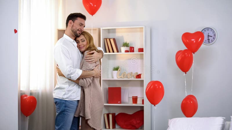 Husband tenderly hugging wife in room with heart-shaped balloons, romantic