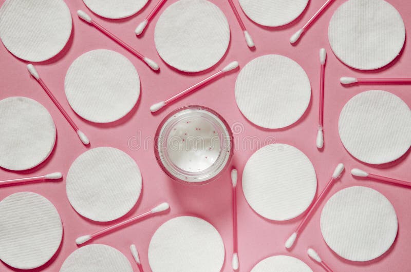 Hygiene products: white round cotton pads and plastic sticks for cleaning the ears lie on a pink background next to face cream