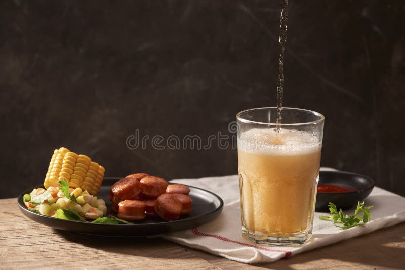 Hällande öl in i exponeringsglas med korven, ketchup, havre och sallad på trätabellen royaltyfri fotografi Hällande öl in i exponeringsglas med korven, ketchup, havre och sallad på trätabellen royaltyfri fotografi