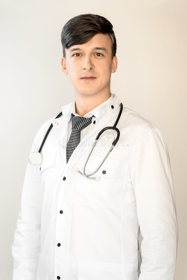 Hälso- och sjukvård En ung läkare med mörkt hår står i en vit kappa med ett stethoscope mot en ljusbakgrund royaltyfri fotografi Hälso- och sjukvård En ung läkare med mörkt hår står i en vit kappa med ett stethoscope mot en ljusbakgrund royaltyfri fotografi