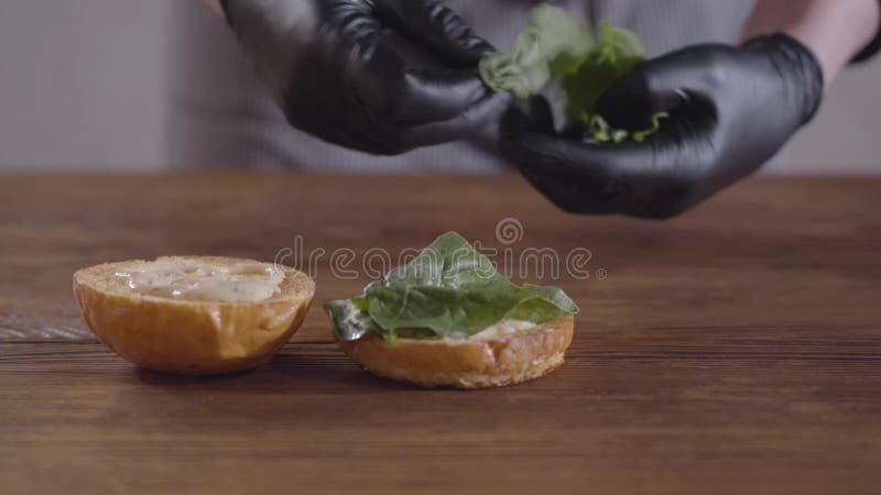 Hände des Chefs in den schwarzen Küchenhandschuhen, die Burgernahaufnahme machen Der Koch, der Basilikumblätter auf den Einteiler