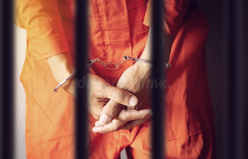 Händer för en fånge i handbojor bak stängerna av ett fängelse i orange jumpsuitkläder arkivbild Händer för en fånge i handbojor bak stängerna av ett fängelse i orange jumpsuitkläder arkivbild