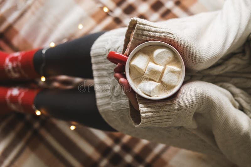 Händer och fot för kvinna` s i tröjan och woolen hemtrevliga röda sockor som rymmer koppen av varmt kaffe med marshmallowen som s royaltyfri foto Händer och fot för kvinna` s i tröjan och woolen hemtrevliga röda sockor som rymmer koppen av varmt kaffe med marshmallowen som s royaltyfri foto