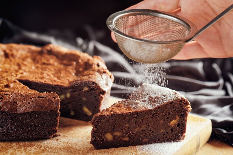 Hands holding sieve with white powdered sugar and sprinkling the powder on piece of freshly baked chocolate brownie cake with walnuts. Homemade sweet dessert on wooden board, family meal. Hands holding sieve with white powdered sugar and sprinkling the powder on piece of freshly baked chocolate brownie cake with walnuts. Homemade sweet dessert on wooden board, family meal.