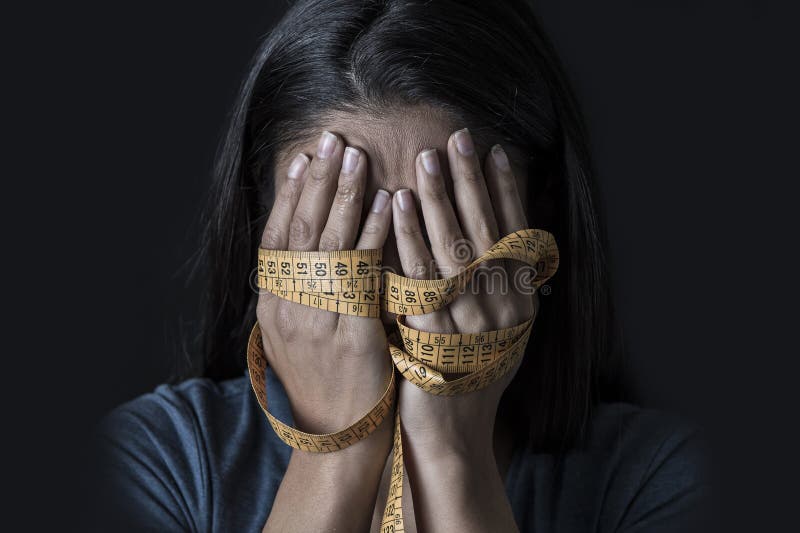 Close up hands wrapped in tailor measure tape covering face of young depressed and worried girl suffering anorexia or bulimia nutrition disorder on black background obsessed with diet and overweight. Close up hands wrapped in tailor measure tape covering face of young depressed and worried girl suffering anorexia or bulimia nutrition disorder on black background obsessed with diet and overweight