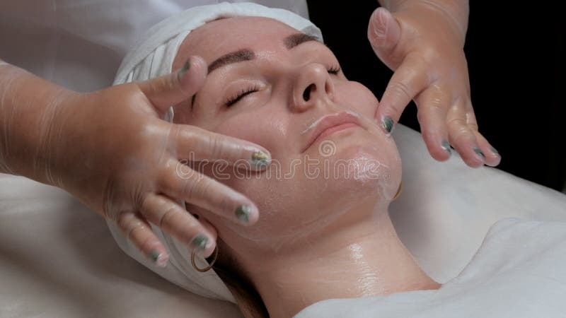 Händerna av cosmetologisten masserar framsidan och applicerar att fukta kräm till de kvinnliga kinderna av klienten Stående av en arkivfoton Händerna av cosmetologisten masserar framsidan och applicerar att fukta kräm till de kvinnliga kinderna av klienten Stående av en arkivfoton