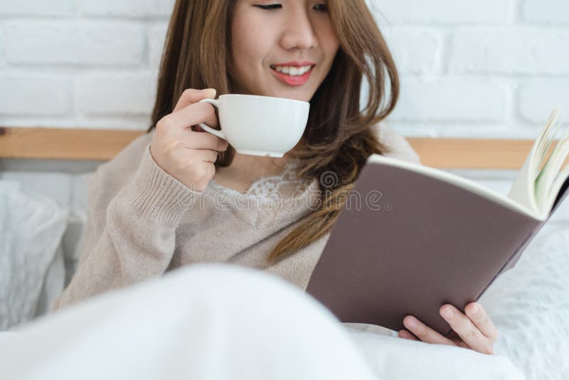 Härlig asiatisk kvinna som tycker om den varma kaffe och läseboken på säng i hennes sovrum royaltyfria bilder