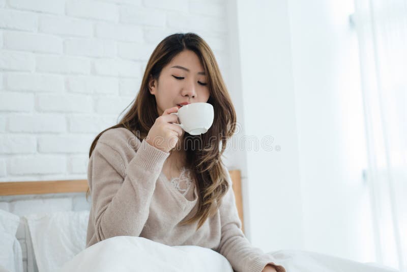 Härlig asiatisk kvinna som tycker om varmt kaffe på säng i hennes sovrum royaltyfri fotografi