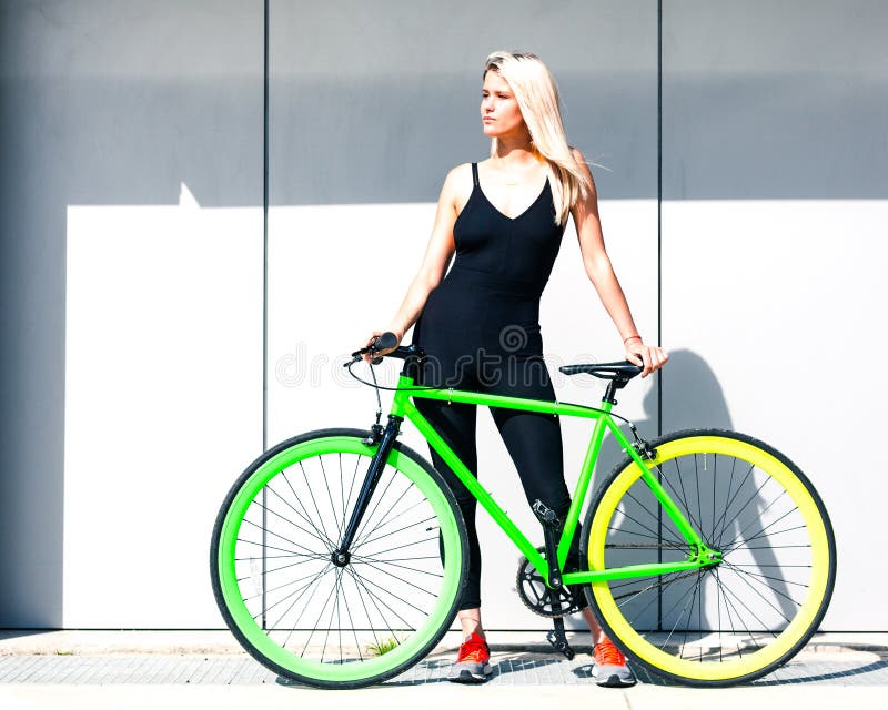 Härlig blond flicka som poserar i sexig svart dräkt på de gråa väggarna av en sommardag med en fast grön cykel arkivfoton