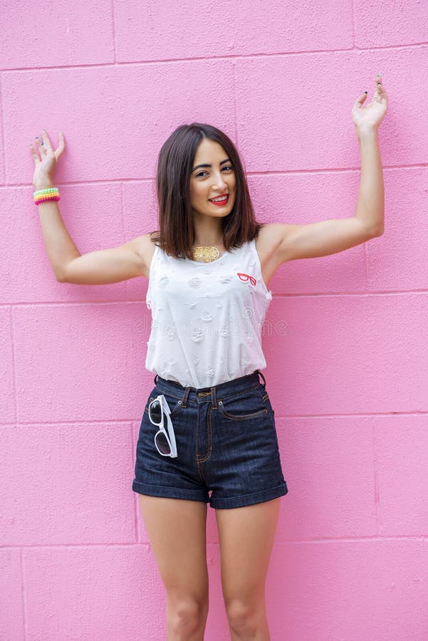 Härlig brunettflicka i vita blus- och jeanskortslutningar som har gyckel på bakgrunden av en rosa vägg som ler, semester royaltyfri fotografi Härlig brunettflicka i vita blus- och jeanskortslutningar som har gyckel på bakgrunden av en rosa vägg som ler, semester royaltyfri fotografi