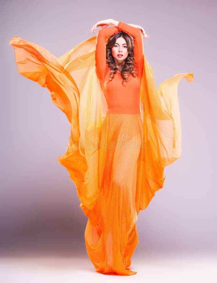 Beautiful woman in long orange dress posing dramatic against grey background. Beautiful woman in long orange dress posing dramatic against grey background