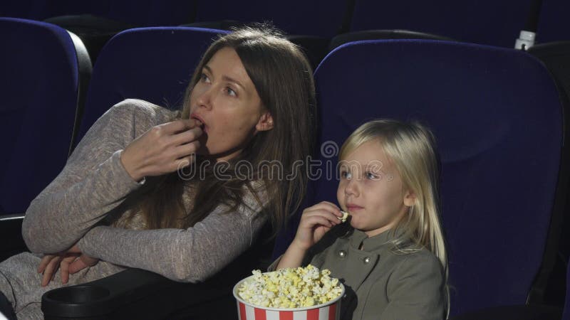 Härlig kvinna och hennes liten dotter som äter hållande ögonen på film för popcorn på bion royaltyfria bilder Härlig kvinna och hennes liten dotter som äter hållande ögonen på film för popcorn på bion royaltyfria bilder