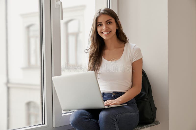 H?rlig kvinnlig student som sitter med b?rbara datorn p? f?nsterbr?da arkivbild H?rlig kvinnlig student som sitter med b?rbara datorn p? f?nsterbr?da arkivbild