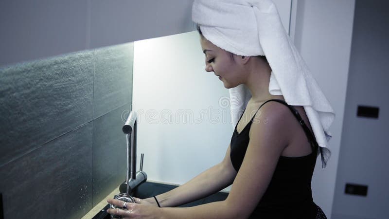 Härlig le ung kvinna med den vita badlakanet på hennes huvud som tvättar ett exponeringsglas i kök Morgonrutin efter dusch