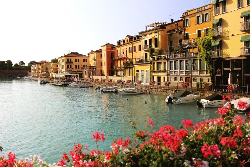 Härlig morgon i den Peschiera del Garda staden Färgrika hus, arkitektursikt med fartyg och suddig pelargon royaltyfria bilder Härlig morgon i den Peschiera del Garda staden Färgrika hus, arkitektursikt med fartyg och suddig pelargon royaltyfria bilder
