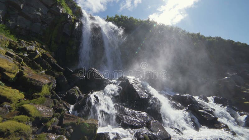 Härlig sikt av vattenfallet Höga klippor som täckas med grön mossa, solen, skiner i ramen, vattendroppar faller på