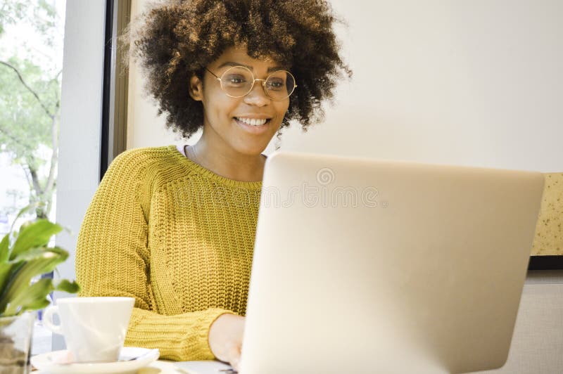 Härlig svart studentkvinna som ler på bärbara datorn som dricker kaffe arkivfoto