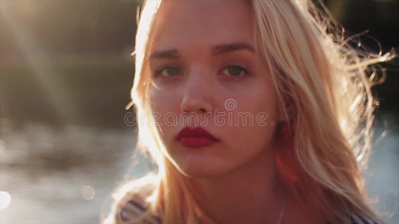 Härlig ung caucasian flicka som framme spelar med hennes blonda hår på solnedgång av vatten och ser in i kameran