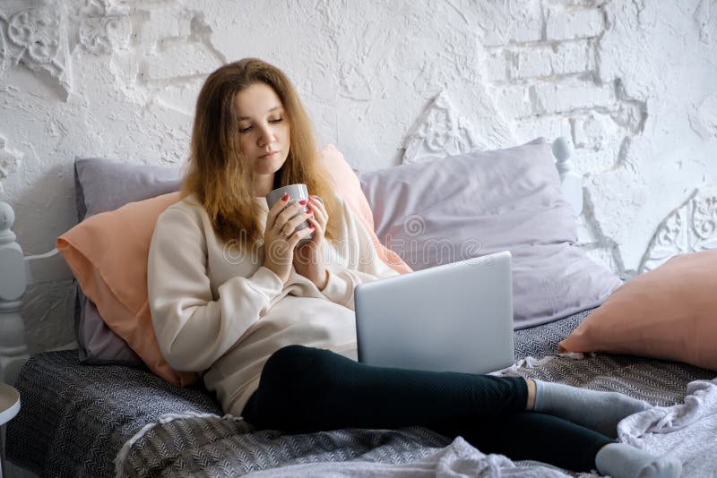 Härlig ung flicka som sitter i morgonen i säng, dricker svart kaffe och ser med intresse i bärbar datorboken arkivfoton Härlig ung flicka som sitter i morgonen i säng, dricker svart kaffe och ser med intresse i bärbar datorboken arkivfoton