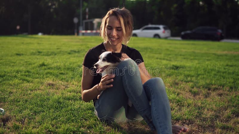 Härlig ung kvinna i tillfälligt sammanträde på gräs med hennes husdjur och le för hund Ägare med den älskvärda lilla hunden utomh