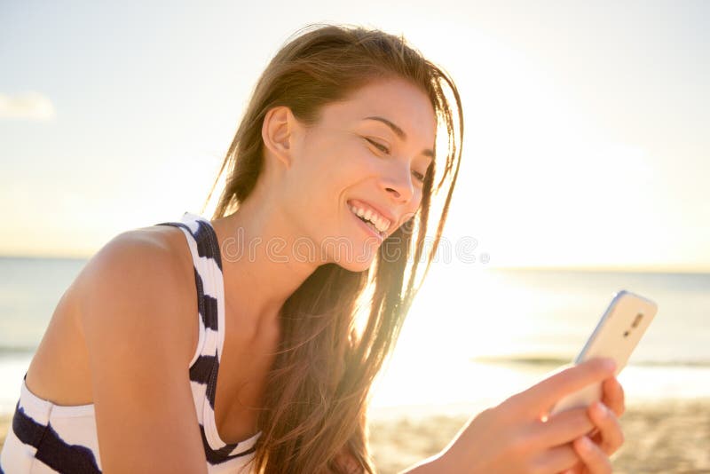 Mixed race pretty smiling young woman holding and looking at smart phone on hawaiian beach in sunset, enjoying summer vacation travel. Sea and horizon in background. Mixed race pretty smiling young woman holding and looking at smart phone on hawaiian beach in sunset, enjoying summer vacation travel. Sea and horizon in background