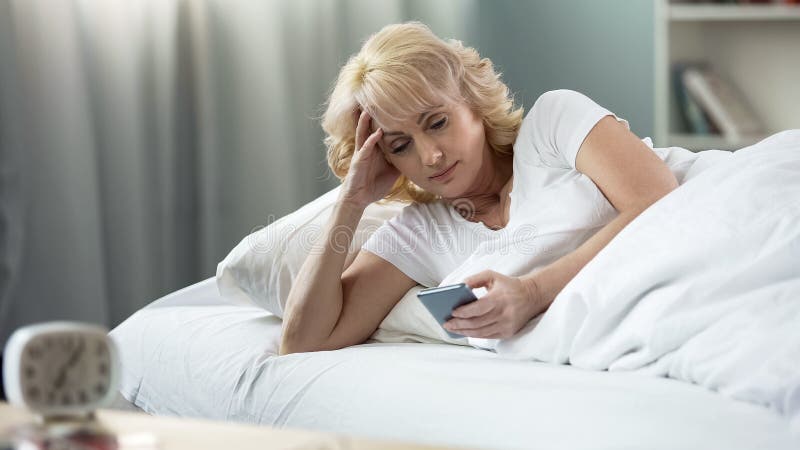 Härlig åldrig kvinna som ligger i säng som pratar i sociala nätverk på smartphonen arkivbild Härlig åldrig kvinna som ligger i säng som pratar i sociala nätverk på smartphonen arkivbild