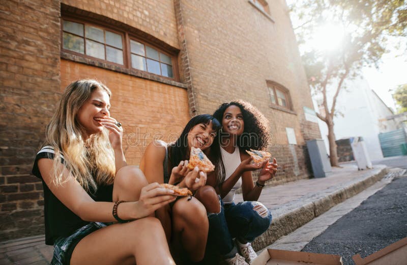 Härliga flickor som utomhus sitter av vägen som äter pizza royaltyfria foton Härliga flickor som utomhus sitter av vägen som äter pizza royaltyfria foton