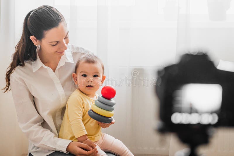 Härligt behandla som ett barn lilla barnet med den unga modern framme av kameran som inomhus vlogging eller blogging arkivbild Härligt behandla som ett barn lilla barnet med den unga modern framme av kameran som inomhus vlogging eller blogging arkivbild