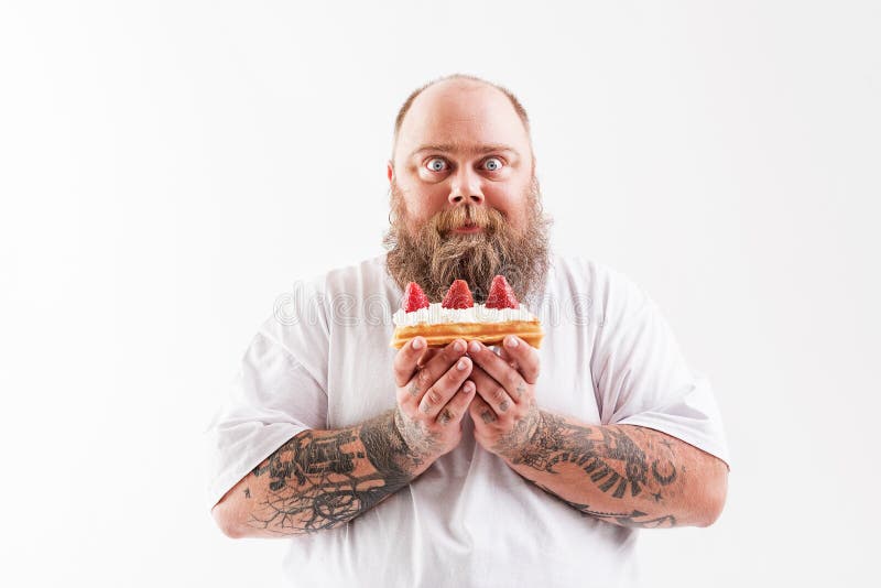 Waist up portrait of happy thick guy eating tasty cake with cream and strawberry. He is standing and staring at camera with desire. Isolated. Waist up portrait of happy thick guy eating tasty cake with cream and strawberry. He is standing and staring at camera with desire. Isolated