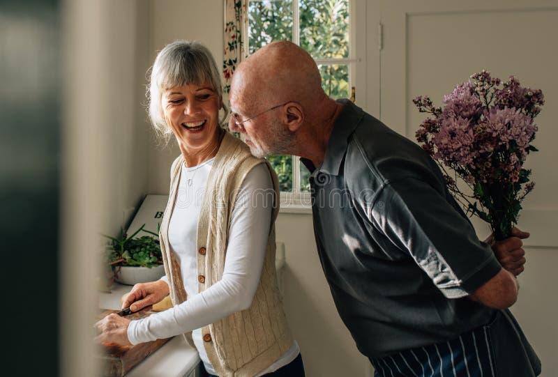 Hög man som döljer en grupp av blommor bak honom för att förvåna hans fru Kvinnaklippgrönsaker i kök medan hennes make arkivbilder