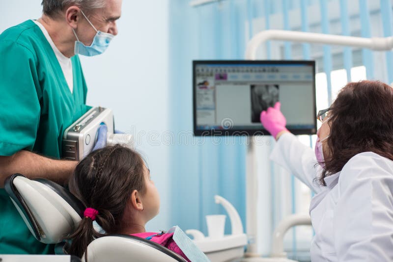 Hög pediatrisk tandläkare med assistenten som gör den tålmodiga flickan för tand- behandling som använder den tand- röntgenappara fotografering för bildbyråer Hög pediatrisk tandläkare med assistenten som gör den tålmodiga flickan för tand- behandling som använder den tand- röntgenappara fotografering för bildbyråer