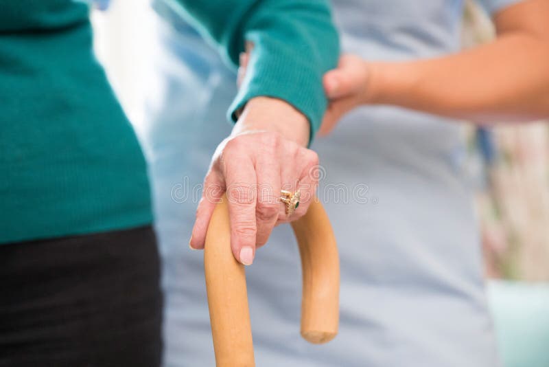 Senior Woman`s Hands On Walking Stick With Care Worker In Background. Senior Woman`s Hands On Walking Stick With Care Worker In Background
