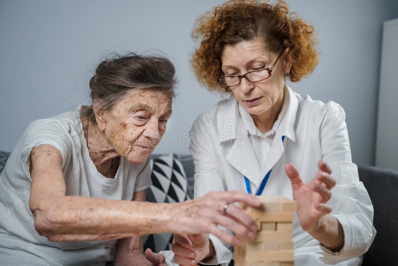 Högre kvinna som spelar Jenga-byggtornet i block. Äldre läkare i vit labbrock som stöder äldre patienter och utvecklar logik. Äldre läkare i vit labblackering som stödjer äldre patienter som utvecklar logiska och fina motoriska färdigheter alzheimsjukdom hemma med gammal ensam kvinna.